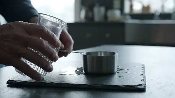A man's hands filling a steel quarter-cup measure with water and tipping it into a small clear glass on a slate counter