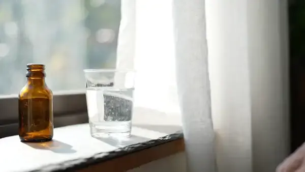 A small glass of water on a sunlit windowsill beside an amber bottle, a hand picking it up at midday as the curtain stirs