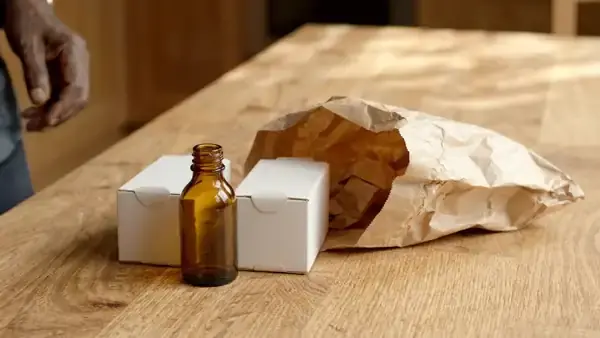 A man's hands placing a small amber bottle and two plain boxes into a brown paper bag on a kitchen table and folding it shut