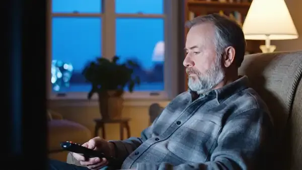 A man in his sixties in a lamp-lit armchair switching off the television and sitting quietly as the room goes still